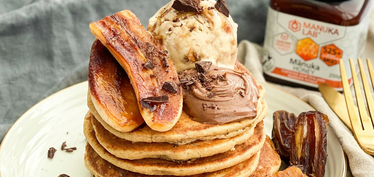A tall stack of fluffy golden pancakes topped with caramelized bananas and dark chocolate shavings, pictured next to a jar of Manuka Lab 525 MGO Manuka Honey.