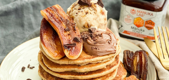 A tall stack of fluffy golden pancakes topped with caramelized bananas and dark chocolate shavings, pictured next to a jar of Manuka Lab 525 MGO Manuka Honey.